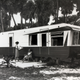 A woman with a clipboard interviews an individual who is seated outside of a mobile home.